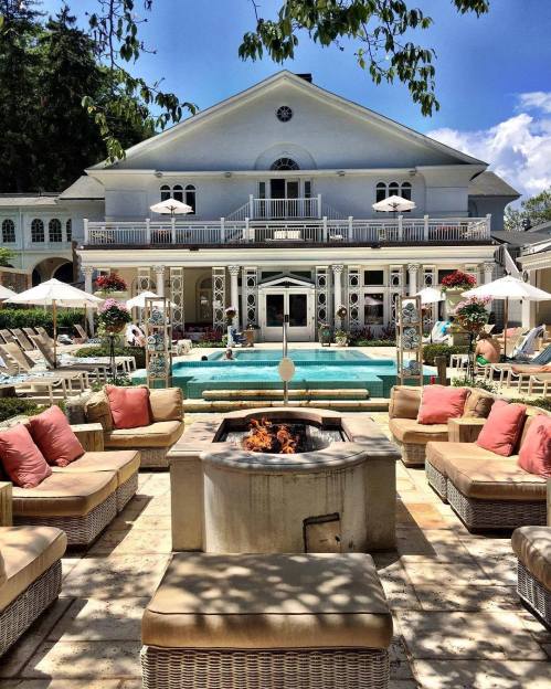 Luxurious outdoor pool area with lounge chairs, fire pit, and a grand house in the background under a clear blue sky.