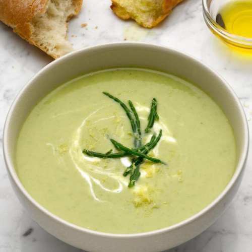 A bowl of green soup garnished with herbs, accompanied by bread and a small dish of olive oil.