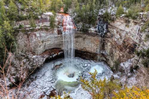 A stunning waterfall cascades into a rocky pool, surrounded by snow-covered cliffs and evergreen trees.