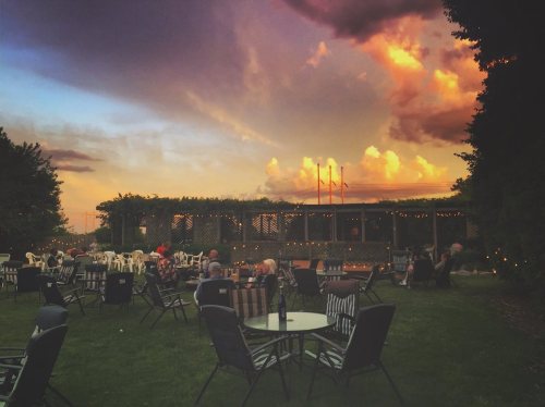 A scenic outdoor venue at sunset, with chairs arranged around tables and colorful clouds in the sky.