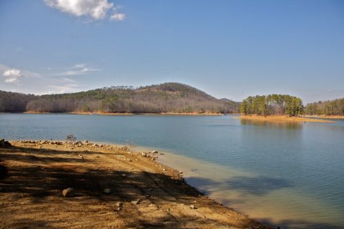 A serene lake surrounded by hills and trees under a clear blue sky, with a sandy shore in the foreground.