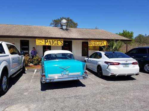 A vintage turquoise car parked next to a white sedan outside Paige's Burgers, with a clear blue sky above.