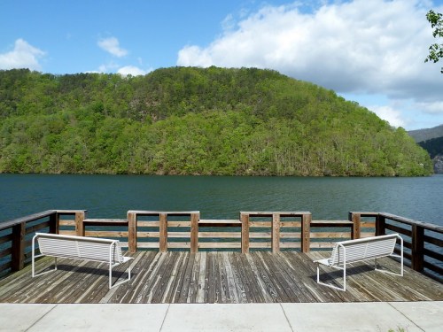 A wooden deck with two benches overlooks a calm lake surrounded by lush green hills under a partly cloudy sky.