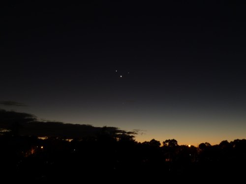 A twilight sky with a gradient from dark to light, featuring three bright celestial objects above a silhouette of trees.