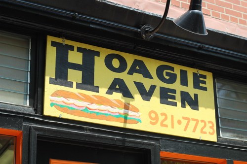 Sign for "Hoagie Haven" featuring a colorful sandwich graphic and contact number, displayed on a storefront.