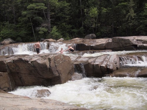 People enjoying a natural rocky river with small waterfalls surrounded by lush greenery.