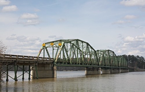 A green metal bridge spans a river, with cloudy skies above and trees lining the riverbank.