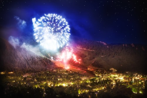 Colorful fireworks illuminate a night sky over a mountainous landscape and a brightly lit town below.