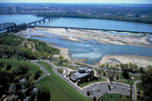 Aerial view of a river with a bridge, dry riverbed, and a park area with a building and parking lot nearby.