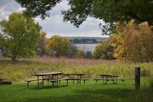 A serene park scene with picnic tables, colorful autumn trees, and a lake in the background under a cloudy sky.