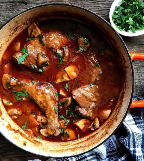 A pot of savory chicken stew with mushrooms and herbs, garnished with fresh parsley on a wooden surface.