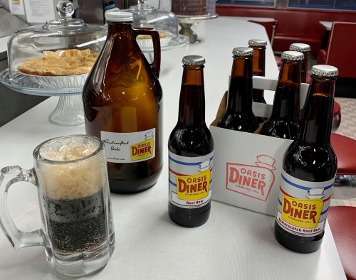 A glass of root beer with froth, a growler, and six bottles of Oasis Diner root beer on a diner counter.