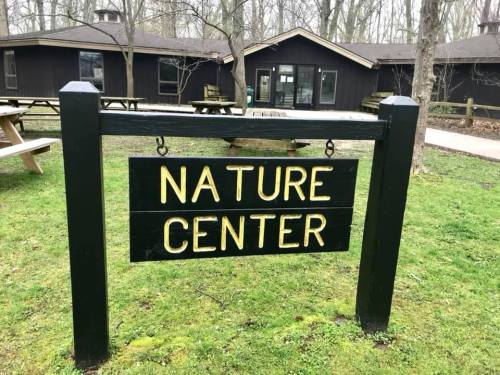 Sign for a Nature Center in a wooded area, with a dark building and picnic tables in the background.
