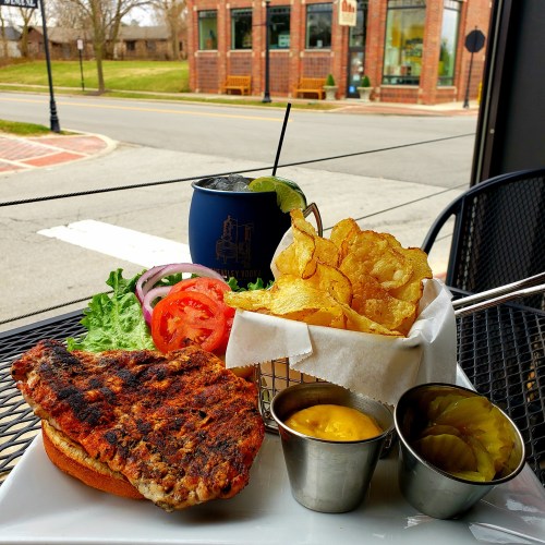 A grilled chicken sandwich with lettuce, tomato, and chips, served with pickles and mustard on a patio table.
