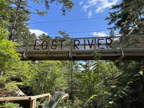A wooden bridge with a sign reading "LOST RIVER" surrounded by lush green trees and a blue sky.