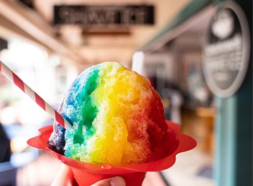 A colorful rainbow snow cone in a red cup, held in front of a blurred café background.