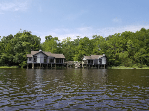Two houses on stilts by a calm lake, surrounded by lush green trees under a clear blue sky.