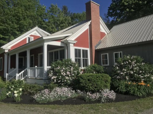 A charming red house with a white porch, surrounded by lush greenery and colorful flower beds under a clear blue sky.