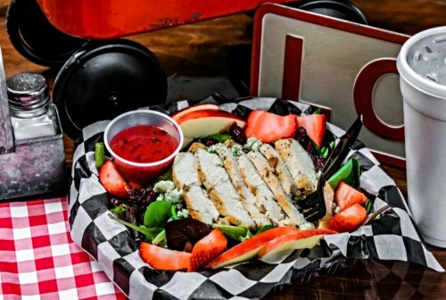 A colorful salad with grilled chicken, apples, and a side of dressing, served in a checkered basket.