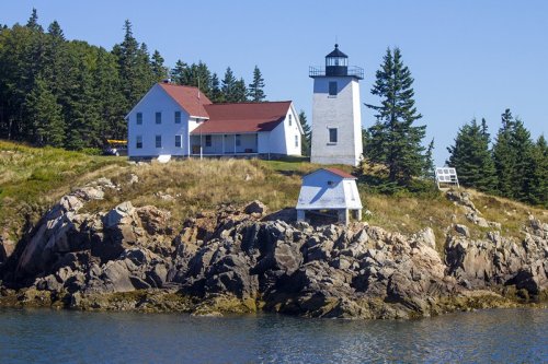 A white lighthouse and house on a rocky shore, surrounded by green trees and blue water under a clear sky.