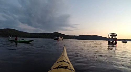 A kayak on calm water at sunset, with another boat and hills in the background under a cloudy sky.
