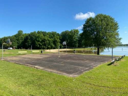 A basketball court beside a lake, surrounded by green grass and trees, with benches in the background.