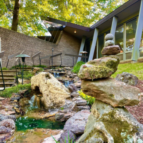 A serene garden scene featuring a stone stack near a small waterfall and a modern building in the background.