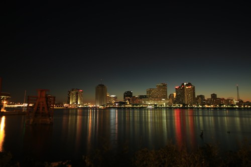 City skyline at night reflecting on water, with illuminated buildings and a clear sky transitioning to dawn.
