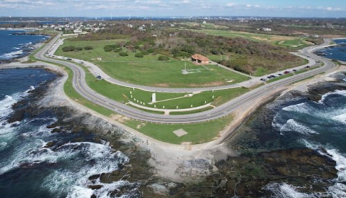 Aerial view of a coastal area with a winding road, green fields, and rocky shoreline under a blue sky.