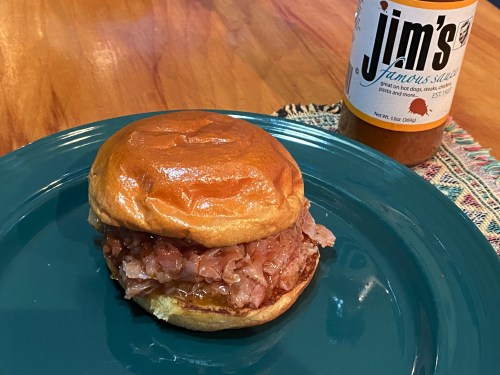 A pulled pork sandwich on a bun, served on a teal plate, with a bottle of Jim's famous sauce in the background.