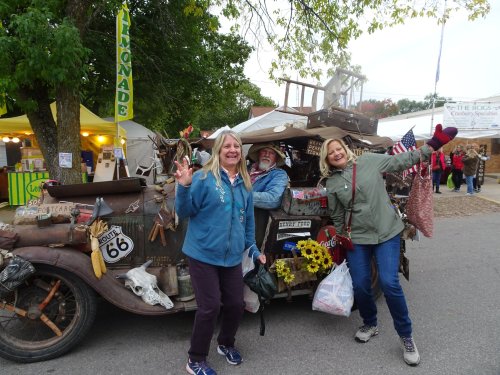 Three smiling people pose in front of a vintage car decorated with various items at a fair.