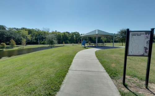 A paved path leads to a gazebo by a pond, with a trail map sign nearby and greenery in the background.