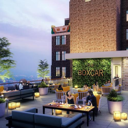 A rooftop terrace with seating, greenery, and a sign reading "THE BOXCAR," set against a city skyline at dusk.