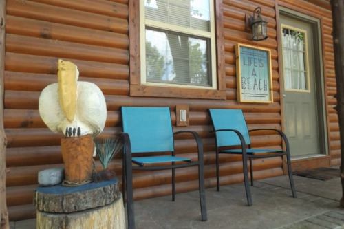 A rustic cabin porch with two blue chairs, a wooden pelican sculpture, and a sign that reads "Life's a Beach."