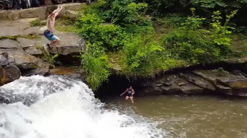 A person jumps off a rock into a river, while another person stands in the water near the shore. Lush greenery surrounds the scene.