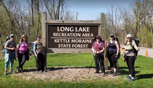 A group of six women with backpacks stands in front of the Long Lake Recreation Area sign in Kettle Moraine State Forest.