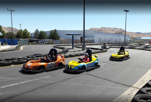 Four go-karts racing on a track, with two drivers in orange and yellow karts and two in black and white karts.