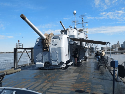 A naval ship deck featuring large cannons, with people exploring and a clear blue sky in the background.
