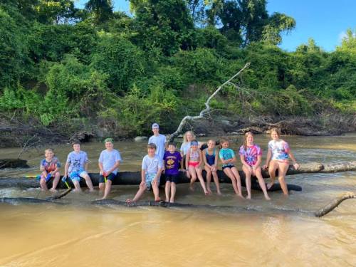 A group of children and adults sitting on a log in a river, surrounded by greenery and enjoying a sunny day.