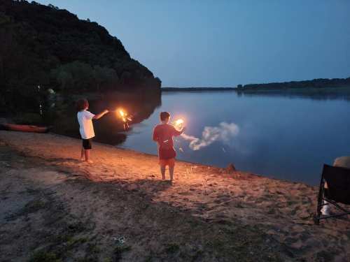 Two children on a beach at dusk, holding sparklers and playing near a calm river.