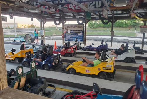 A go-kart track with several colorful karts lined up, and drivers preparing to race in a sunny outdoor setting.