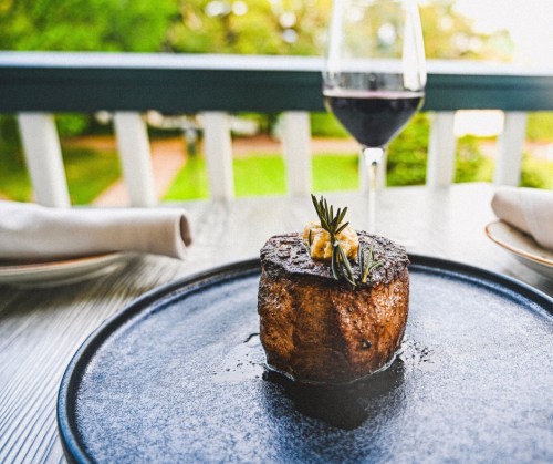 A perfectly cooked steak garnished with herbs, served on a black plate with a glass of red wine in the background.