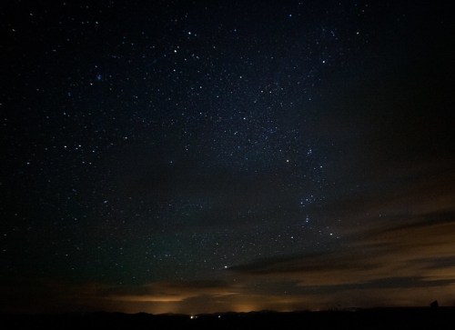 A starry night sky filled with twinkling stars and wispy clouds, with faint lights on the horizon.