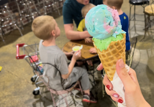 A hand holds a colorful ice cream cone, with children sitting at a table in the background.