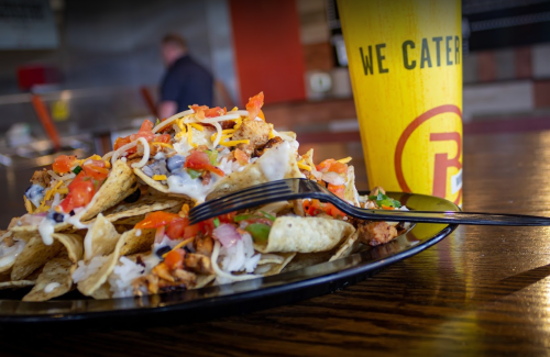 A plate of nachos topped with cheese, tomatoes, and chicken, alongside a drink in a "We Cater" cup.