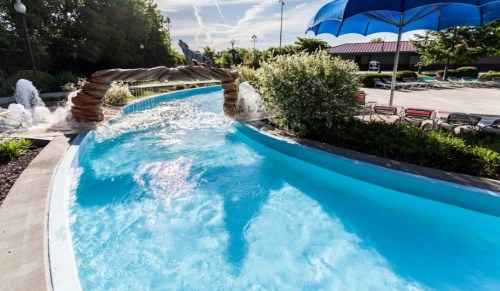A serene pool area featuring a winding lazy river, lush greenery, and a blue umbrella in the background.
