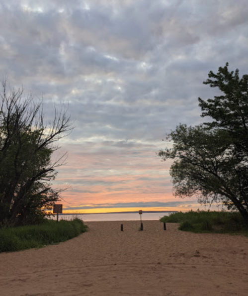 A sandy path leads to a calm beach at sunset, framed by trees and a cloudy sky.