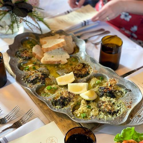 A platter of baked escargot with lemon wedges and bread, set on a table with utensils and a menu in the background.