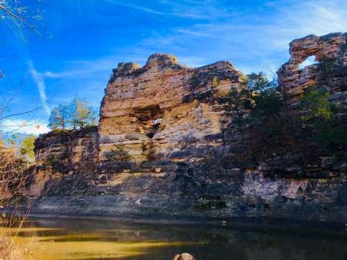 Rocky cliffside with a natural arch, surrounded by trees and reflecting in a calm river under a blue sky.