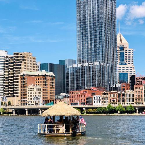 A small boat with a thatched roof floats on a river, surrounded by a city skyline of tall buildings.
